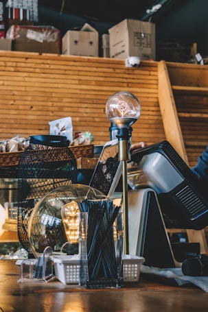 A cozy interior featuring a wooden counter with a decorative lamp. The counter displays various items, including a basket with a netting pattern, a transparent box with black sticks, and a glowing globe light. In the background, cardboard boxes are stacked against a wooden paneled wall.