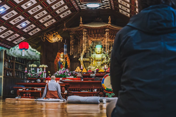 A serene temple setting with a large golden Buddha statue at the center, surrounded by vibrant flowers and offerings. A monk in traditional robes is seated on the floor, facing the altar, while another person is visible in the foreground. The ceiling is ornately decorated with patterned tiles, and a red lantern hangs to the side.