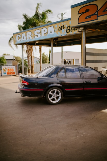 A black car with red detailing is parked at an outdoor car wash facility. The facility has a sign reading 'CAR SPA' and another area advertising a 'DOG WASH'. Palm trees and buildings are visible in the background. The car appears to be an older model sedan, and the setting gives a slightly nostalgic vibe.