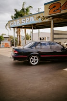 A black car with red detailing is parked at an outdoor car wash facility. The facility has a sign reading 'CAR SPA' and another area advertising a 'DOG WASH'. Palm trees and buildings are visible in the background. The car appears to be an older model sedan, and the setting gives a slightly nostalgic vibe.