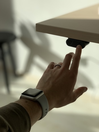 A hand wearing a smartwatch is seen pointing towards a button under a wooden desk. The lighting creates shadows on the light-colored background, suggesting a minimalist and modern setting.