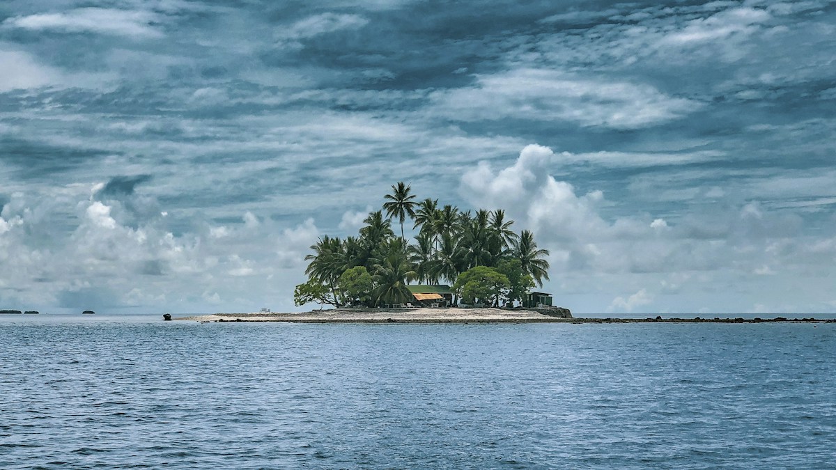 Petite île couverte de cocotiers posée sur une mer calme, sous un ciel chargé de nuages