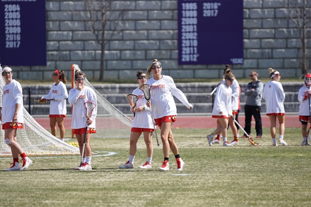 A group of athletes wearing white and red uniforms are gathered on a grassy field with lacrosse sticks. Some are standing near a goal and others are walking around. A stone wall and a scoreboard with dates are visible in the background. The scene appears to be during practice or a warm-up session.
