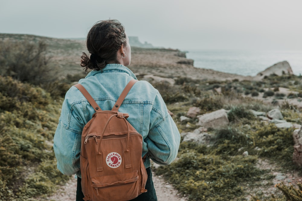 woman wearing blue denim jacket with brown backpack walking on trail near beach