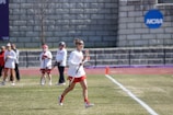 A group of female athletes are on a grassy sports field. One is in the foreground running with a lacrosse stick, wearing a white jersey and red shorts. Other players and possibly coaches are in the background, some holding lacrosse sticks. The field has white markings and a purple track nearby. In the background, there is a grey stone wall with a blue NCAA sign.