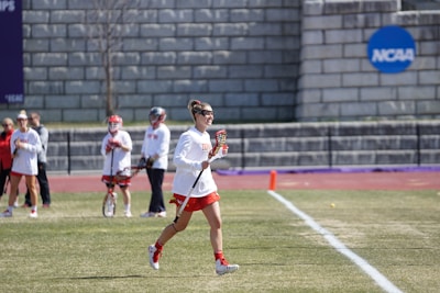 A group of female athletes are on a grassy sports field. One is in the foreground running with a lacrosse stick, wearing a white jersey and red shorts. Other players and possibly coaches are in the background, some holding lacrosse sticks. The field has white markings and a purple track nearby. In the background, there is a grey stone wall with a blue NCAA sign.