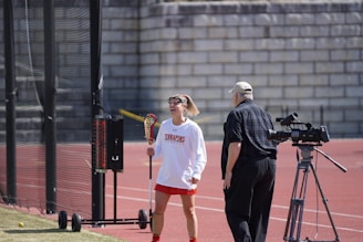 A female lacrosse player in athletic attire and headband is standing on a sports field, holding a lacrosse stick with a ball in the netting, while smiling and interacting with an older man who is operating a video camera on a tripod. The background includes a fencing and a brick wall.