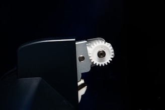 A close-up view of a mechanical component featuring a white gear partially enclosed within a dark metallic or plastic casing. The gear is attached to a metal structure, with bolts visible near the center of the gear.