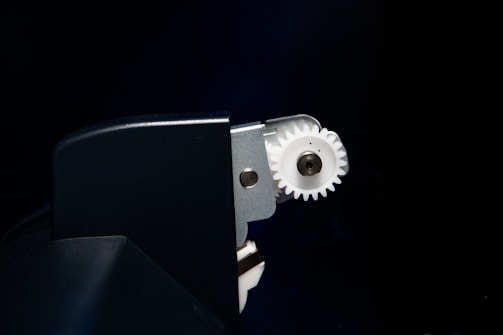 A close-up view of a mechanical component featuring a white gear partially enclosed within a dark metallic or plastic casing. The gear is attached to a metal structure, with bolts visible near the center of the gear.
