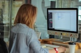 A person with brown hair is seated at a desk, working on a computer. The monitor displays a file directory, and the computer is adorned with colorful stickers depicting various emoji and icons. The desk also holds a coffee cup and a calendar displaying the month of October. The person is wearing a blue and white striped shirt.