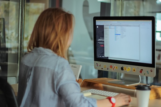 A person with brown hair is seated at a desk, working on a computer. The monitor displays a file directory, and the computer is adorned with colorful stickers depicting various emoji and icons. The desk also holds a coffee cup and a calendar displaying the month of October. The person is wearing a blue and white striped shirt.