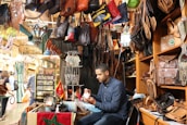 A man sits inside a market stall filled with leather bags and goods. Various bags hang from the ceiling, and shelves are lined with intricately designed items. The setting appears busy, with people walking by outside the stall. Bright, decorative lanterns hang overhead, adding warmth to the scene.