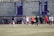 A sports team is practicing or playing on a grassy field in front of a large stone wall with banners displaying men's soccer achievements. People are wearing sports attire, and some are engaged in conversation or coaching.