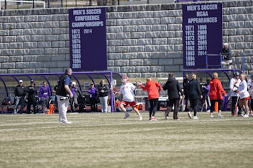 A sports team is practicing or playing on a grassy field in front of a large stone wall with banners displaying men's soccer achievements. People are wearing sports attire, and some are engaged in conversation or coaching.
