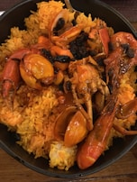 A dish consisting of yellow rice topped with a variety of seafood, including prawns and crab, served in a black bowl. The seafood is coated in a rich, red sauce and garnished with black olives or raisins.
