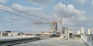 A modern train is located at an elevated railway track with overhead lines, set against a cityscape backdrop. The sky is partially cloudy and several tall buildings are visible in the distance.
