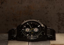 A black wristwatch with a dark dial and metallic band is placed on a textured surface. The watch features several small dials and bold hour markers, with light reflecting off its glossy surface. The background has an industrial, rustic appearance with uneven tones.