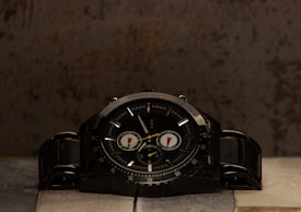 A black wristwatch with a dark dial and metallic band is placed on a textured surface. The watch features several small dials and bold hour markers, with light reflecting off its glossy surface. The background has an industrial, rustic appearance with uneven tones.