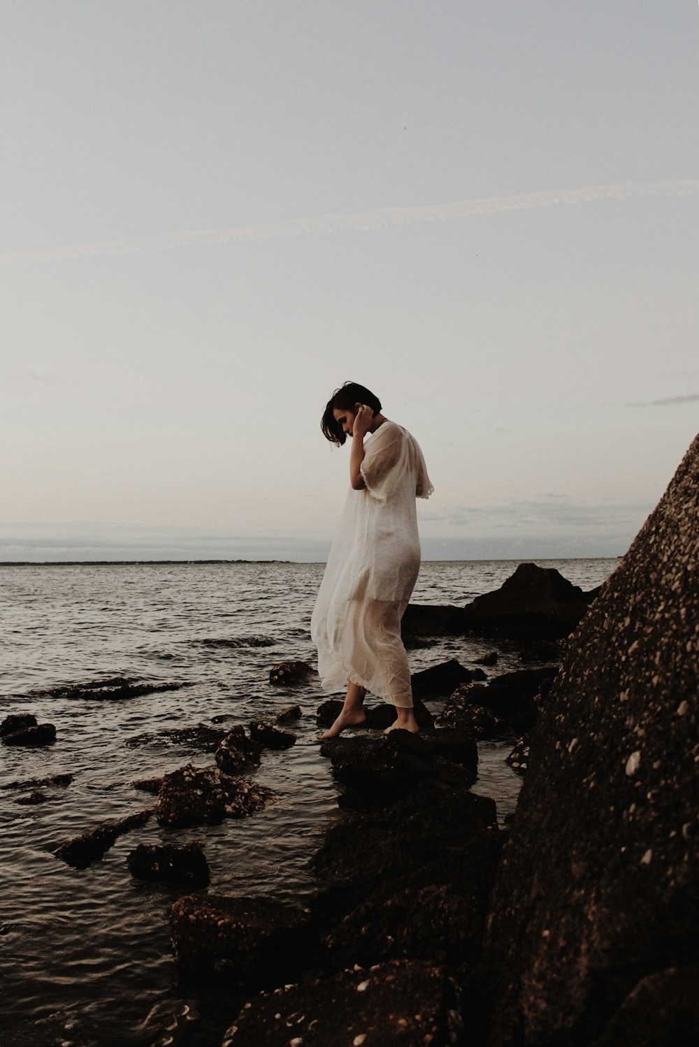 흰 얇은 드레스를 입은 여자가 해변가의 바위 위를 걷고 있습니다