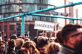 A crowd of people gathered in an urban area, with a prominent sign reading 'DAGEGEN!' held up above their heads. Modern buildings serve as a backdrop.