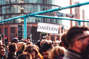 A crowd of people gathered in an urban area, with a prominent sign reading 'DAGEGEN!' held up above their heads. Modern buildings serve as a backdrop.