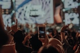 A crowded concert setting captured with an audience holding up a smartphone to record the live performance. Bright stage lights create a vibrant backdrop, while the crowd engages enthusiastically, with some raising their hands in the air.