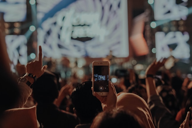 A crowded concert setting captured with an audience holding up a smartphone to record the live performance. Bright stage lights create a vibrant backdrop, while the crowd engages enthusiastically, with some raising their hands in the air.