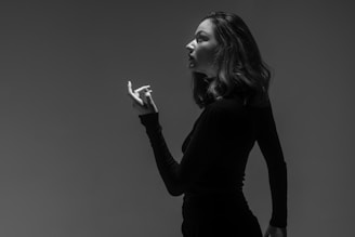 A monochrome photograph of a woman in profile, standing against a plain background. The woman is wearing a black long-sleeve top and her hair is styled in loose waves. Her expression appears contemplative and her hand is raised slightly with fingers elegantly poised.