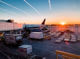 A serene airport scene at sunset with an airplane parked near the terminal gate. Luggage carts and utility vehicles are spread across the tarmac. The sky displays a gradient of orange to blue as the sun sets on the horizon, casting a warm glow over the area.
