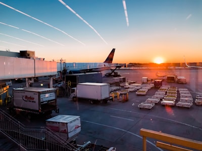 A serene airport scene at sunset with an airplane parked near the terminal gate. Luggage carts and utility vehicles are spread across the tarmac. The sky displays a gradient of orange to blue as the sun sets on the horizon, casting a warm glow over the area.