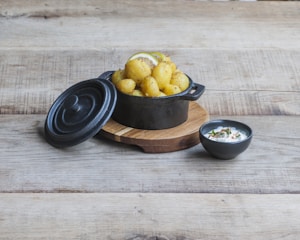 A rustic wooden table displays a cast iron pot filled with seasoned potatoes, garnished with lemon slices. The lid is slightly tilted to the side. Next to the pot, a small black bowl contains a dip topped with herbs and spices.