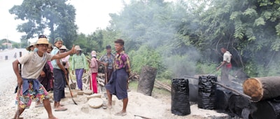 A group of people are engaged in an outdoor activity that appears to involve making charcoal or some other form of manual labor. Smoke rises from several blackened barrels, and logs lie around on the ground. These individuals are casually dressed, some with hats, in a rural setting surrounded by trees.