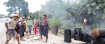 A group of people are engaged in an outdoor activity that appears to involve making charcoal or some other form of manual labor. Smoke rises from several blackened barrels, and logs lie around on the ground. These individuals are casually dressed, some with hats, in a rural setting surrounded by trees.