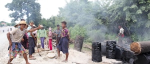 A group of people are engaged in an outdoor activity that appears to involve making charcoal or some other form of manual labor. Smoke rises from several blackened barrels, and logs lie around on the ground. These individuals are casually dressed, some with hats, in a rural setting surrounded by trees.