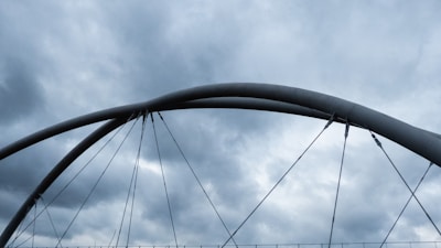 A large arch bridge structure is silhouetted against a cloudy sky. The curved metal beams are supported by cables, creating a modern architectural design.