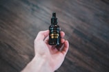 A hand is holding a dark glass bottle with a dropper lid. The label on the bottle reads 'Zennutt Beard Oil.' The background consists of a blurred, dark wooden surface.