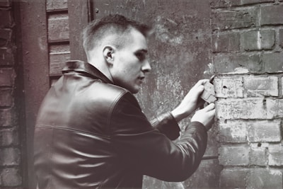 A person wearing a leather jacket is using a tool to tamper with a brick wall in an outdoor setting. The scene has a gritty, urban feel with worn bricks and a metallic door visible in the background.
