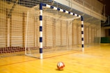 A handball goalpost with a blue and white pattern stands prominently on a wooden indoor court. The netting is visible, and a handball lies on the floor in front of the goal.
