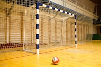 A handball goalpost with a blue and white pattern stands prominently on a wooden indoor court. The netting is visible, and a handball lies on the floor in front of the goal.