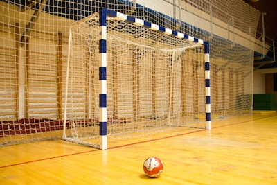 A handball goalpost with a blue and white pattern stands prominently on a wooden indoor court. The netting is visible, and a handball lies on the floor in front of the goal.