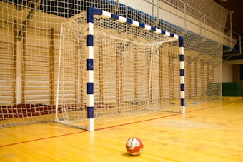 A handball goalpost with a blue and white pattern stands prominently on a wooden indoor court. The netting is visible, and a handball lies on the floor in front of the goal.