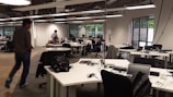 An open-plan office with multiple workstations and people working at computers. The room has modern, minimalist design with white desks, black chairs, and large windows letting in natural light. Some workers are focused on their screens while one person walks across the room.