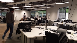An open-plan office with multiple workstations and people working at computers. The room has modern, minimalist design with white desks, black chairs, and large windows letting in natural light. Some workers are focused on their screens while one person walks across the room.