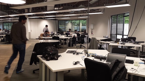 An open-plan office with multiple workstations and people working at computers. The room has modern, minimalist design with white desks, black chairs, and large windows letting in natural light. Some workers are focused on their screens while one person walks across the room.