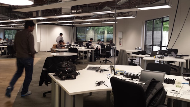 An open-plan office with multiple workstations and people working at computers. The room has modern, minimalist design with white desks, black chairs, and large windows letting in natural light. Some workers are focused on their screens while one person walks across the room.