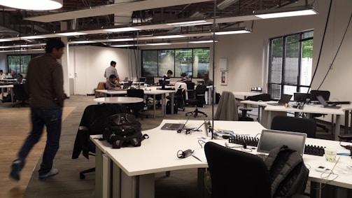 An open-plan office with multiple workstations and people working at computers. The room has modern, minimalist design with white desks, black chairs, and large windows letting in natural light. Some workers are focused on their screens while one person walks across the room.