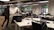 An open-plan office with multiple workstations and people working at computers. The room has modern, minimalist design with white desks, black chairs, and large windows letting in natural light. Some workers are focused on their screens while one person walks across the room.