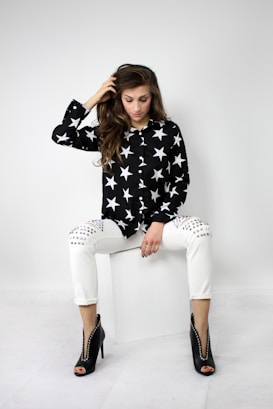 A stylish person is sitting on a white cube. They are wearing a black shirt with white star patterns, white studded pants, and black high-heeled shoes with circular cutouts. Their long hair is styled in loose waves and they are gazing down, with one hand gently placed on their head.