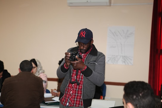 A person in a cap is focusing on a camera, appearing engaged in taking a photograph. There are several people sitting at tables, and some papers are scattered around. A large poster with tree-like illustrations is visible on the wall.