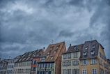 An array of traditional European buildings with steeply pitched roofs under a dramatic, cloudy sky. The architecture features a variety of colors, including cream, blue, and brown tones. Windows are prominent, with some shutters visible, adding to the historic charm.